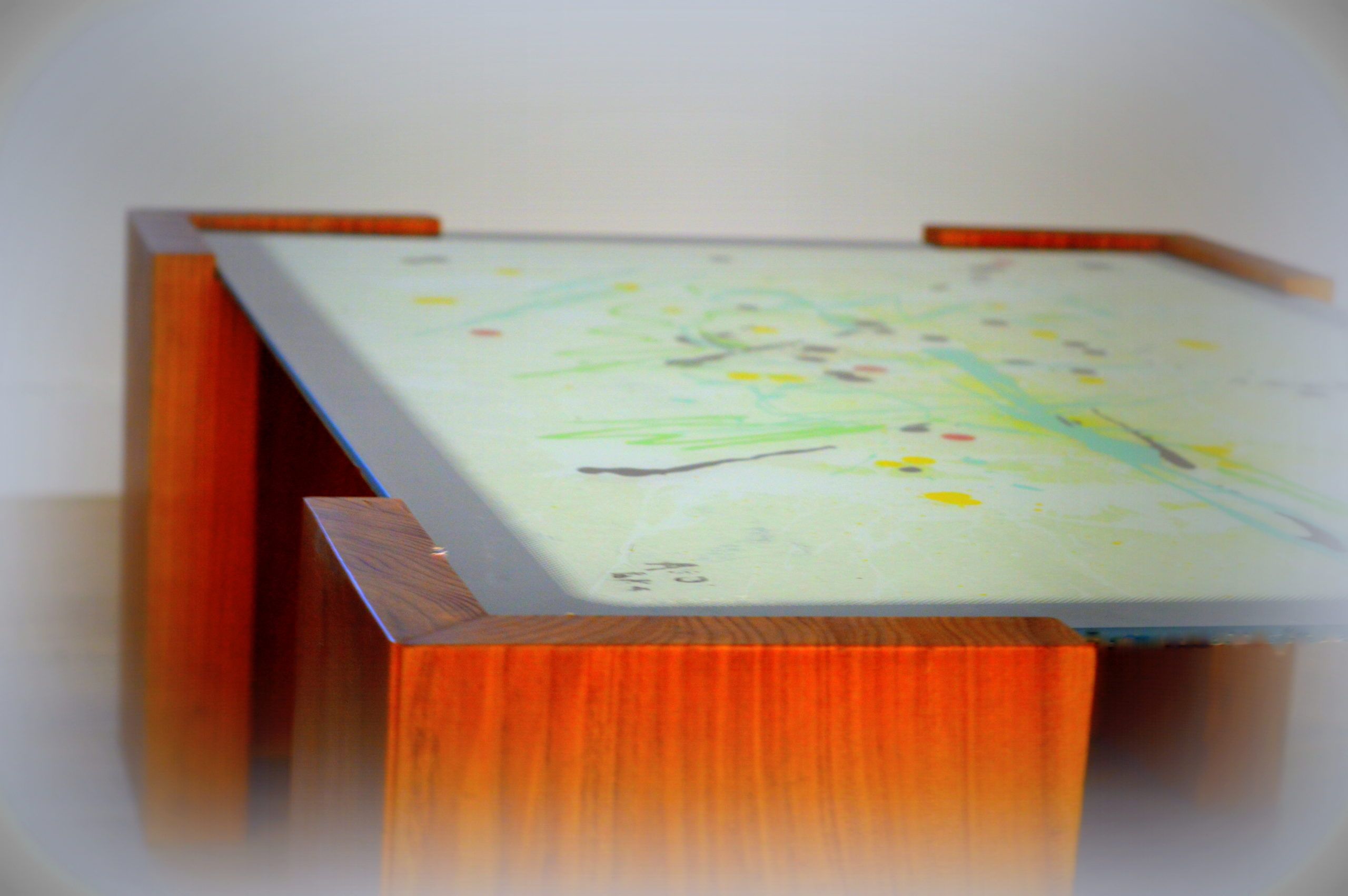 Low wooden table whose glass top shows an abstract work with green lines and yellow, red and grey dots