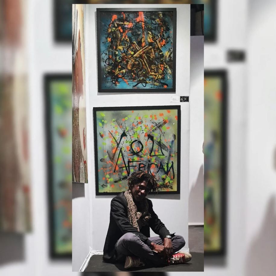 Akoi AKA sitting in front of two of his paintings at an exhibition